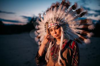 Portrait of young American Indian woman on sunset. Cherokee, Navajo, west native culture. Headdress made of feathers of wild birds. 