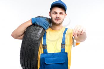 Tyre service, worker in blue uniform holds tire and car keys in hands, white background, repairman, wheel mounting
