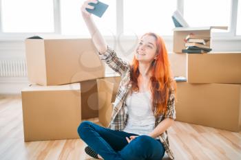 Smiling woman sitting on the floor among cardboard boxes and makes selfie on mobile phone camera, housewarming. Relocation to new home