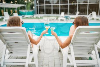 Two sexy girls with cocktails lies on deck chairs near the swimmimg pool. Slim women sitting by the poolside, resort holidays 