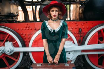 Woman in red hat with suitcase in hands against vintage steam locomotive, red wheels closeup. Old train. Railway engine, railroad journey 