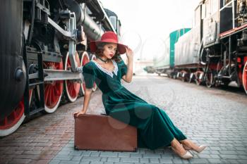 Young old-fashioned woman sitting on suitcase against vintage steam train, red wheels closeup. Old locomotive. Railway engine, travel by railroad