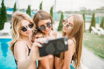 Three sexy girls in swimsuits and sunglasses makes selfie near the swimming pool. Resort holidays. Tanned women on summer holidays