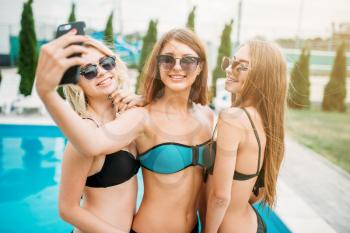 Three sexy girls in swimsuits and sunglasses makes selfie near the swimming pool. Resort holidays. Tanned women on summer holidays