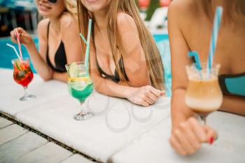 Three sexy female persons in swimsuits drinks cocktails on the poolside. Resort holidays. Tanned women relax in the swimmimg pool