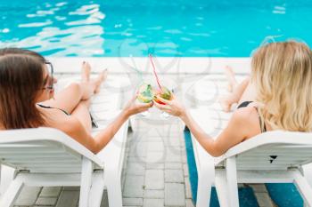Two sexy girls relax with cocktails on deck chairs near the swimmimg pool. Slim women sitting by the poolside, resort holidays