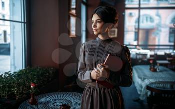Young female person stands near window with book in her hands. Retro style lady in vintage cafe. Brunette young woman in vintage dress dark gray color.