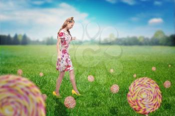 Young pretty girl walking on field with grass and colorful candies. Bright girl with blonde curly hair. Stylish girl in colorful summer dress and yellow shoes, summer meadow on background.