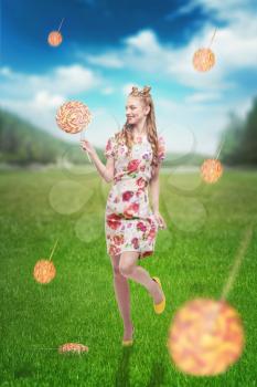 Young pretty girl stands on field with grass and colorful flying lollipops. Bright girl with blonde curly hair. Stylish girl in colorful summer dress and yellow shoes, summer meadow on background.