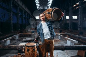 Young male drummer holds wooden drum on the shoulder, factory shop on background. Djembe, musical percussion instrument,