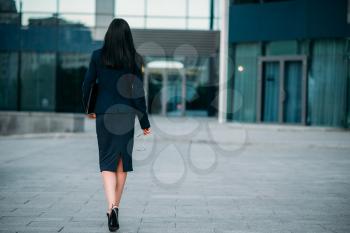 Business woman with eyeglasses and laptop in hands, back view. Modern building, financial center, cityscape. Successful female businessperson