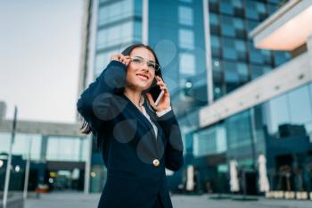 Businesswoman in suit talks by mobile phone outdoor, business center on background. Successful female businessperson