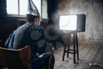 Afraided man in aluminum foil helmet sits in chair and watches tv, paranoia concept. UFO, conspiracy theory, brain theft protection, phobia