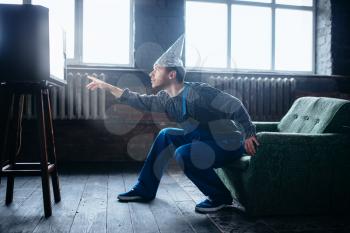 Strange man in tinfoil cap reaches out to the TV, paranoia concept. UFO, conspiracy theory, brain theft protection, phobia