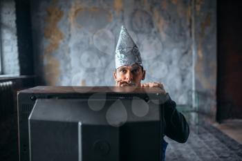 Afraid man in aluminum foil cap watch TV, mind protection, paranoia concept. UFO, conspiracy theory, telepathy phobia