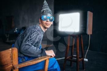 Strange man in tinfoil helmet and hypnotizing glasses watch TV, mind protection, paranoia concept. UFO, conspiracy theory, telepathy phobia