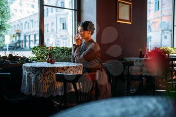 Young beautiful retro lady in cafe sitting near window. Beautiful young girl resting in a cafe and smiling. Dark hair woman looking in window in vintage style restaurant.