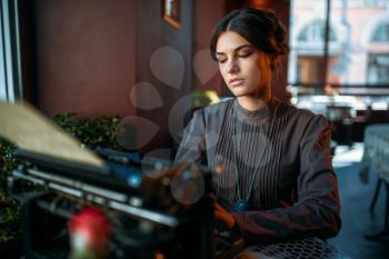 Serious dark hair lady prints on ancient typewriter in cafe. Portrait of retro style woman in vintage cafe prints, close up shot. Brunette woman in dark dress, old style jewelry.