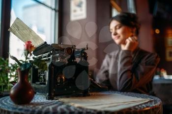 Smiling dark hair lady sits by table with ancient typewriter. Portrait of retro style woman in vintage cafe with typewriter close up shot. Retro style cafe, vase with rose on a table.