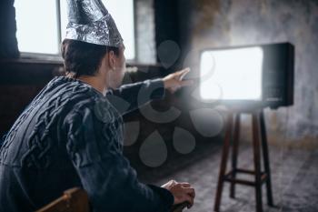Afraided man in aluminum foil helmet reaches out to the TV, paranoia concept. UFO, conspiracy theory, brain theft protection, phobia