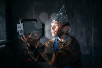 Paranoid man in tinfoil cap watch TV, mind protection from telepathy, paranoia concept. UFO phobia, conspiracy theory
