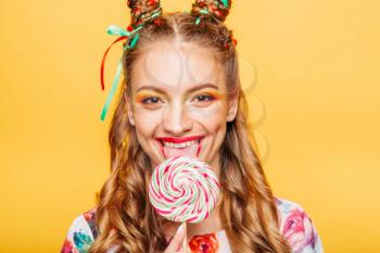 Beautiful young woman with playful look lick candy with tongue. Stylish girl with blonde curly hair. Portrait of attractive lady with big lollypop, yellow wall on background.