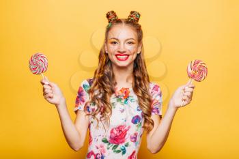 Young smiling lady holding two huge colorful lollypops. Amasing young woman fills happy and holding big candies in her hands. Stylish girl in summer dress, yellow wall on background.