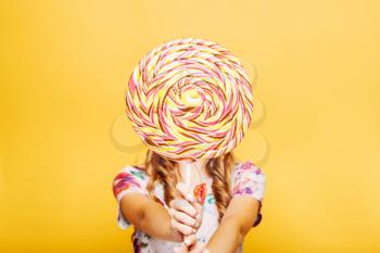 Beautiful young woman with huge candy instead of a head. Big colorful caramel lollypop in hands of woman. Portrait of attractive lady with big lollypop, yellow wall on background.