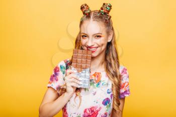 Beautiful young woman with playful look bites huge chocolate. Stylish girl with blonde curly hair. Stylish girl in colorful summer dress, yellow wall on background.
