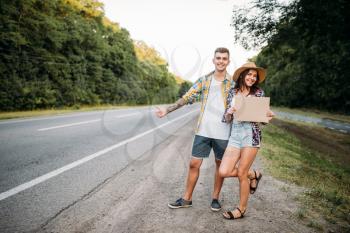 Hitchhiking couple holds blank cardboard with copy space. Hitchhike adventure of young man and woman. Happy hitchhikers on road