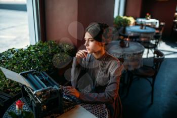 Portrait in profile of beautiful young woman in cafe with ancient typewriter. Woman smiling and prints something in vintage cafe. Dark hair girl sits by the table in restaurant.