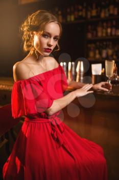 Portrait of young woman in red dress standing at the bar counter. Beautiful lady with beverage in hand in club