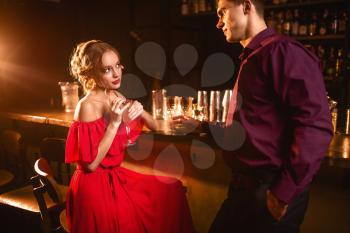 Young woman in red dress flirts with man behind bar counter. Date in nightclub, attractive love couple in pub