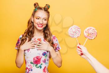 Beautiful young woman surprised somebody gives her lollypops. Bright girl with blonde curly hair. Stylish girl in summer colorful dress, yellow wall on background.