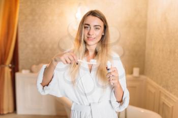 Young woman in white bathrobe brushing her teeth with toothbrush, bathroom interior on background. Morning mouth hygiene, tooth cleaning