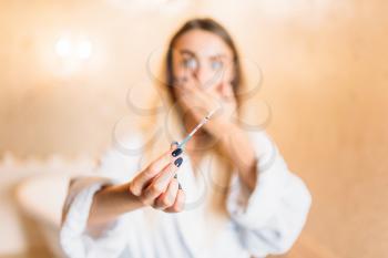 Frightened young woman in white bathrobe with positive pregnancy test, bathroom interior on background. Bodycare and hygiene, healthcare 