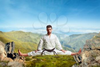 Martial arts karate master in white kimono and black belt doing extreme stretching exercise between the tops of the mountains
