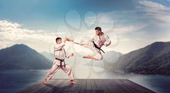Martial arts karate masters in white kimono and black belts, outdoors practice, kick in jump, training on the pier