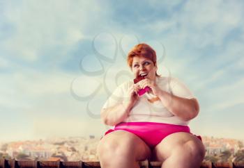 Overweight woman sits on the roof and eats sweet cake, laziness and obesity, bulimic. Unhealthy food eating, fatty female