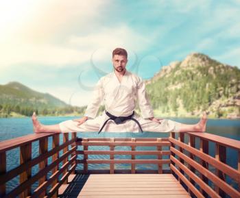 Martial arts karate master in white kimono and black belt doing stretching exercise on the railings of the pier, outdoor training