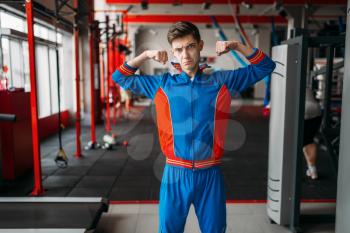Skinny man in sportswear shows his muscles at the gym, humor. Weak male person in sport club