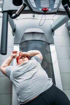 Fat tired woman lies on a treadmill after running in gym, top view. Calories burning, obese female person workout in sport club