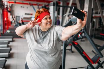 Fat sweaty woman makes selfie against exercise machines in gym. Calories burning, obese female person in sport club