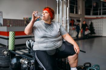 Fat sweaty woman drinks water after active training in gym. Calories burning, obese female person on workout in sport club