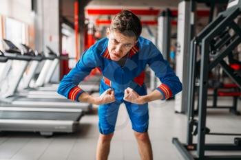 Skinny man in sportswear shows his muscles at the gym, humor. Weak male person in sport club