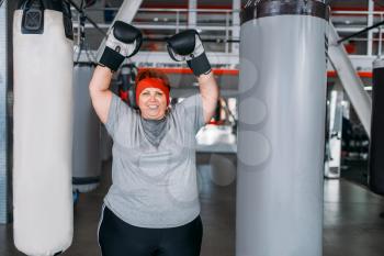 Overweight sweaty woman in boxing gloves against punching bag in gym. Calories burning, obese female person on hard training in sport club