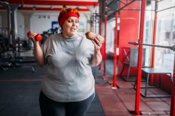 Fat woman with fast food and dumbbell in hands, motivation, hard workout in gym. Calories burning concept, obese female person in fitness club, fat-burning, sport against unhealthy food
