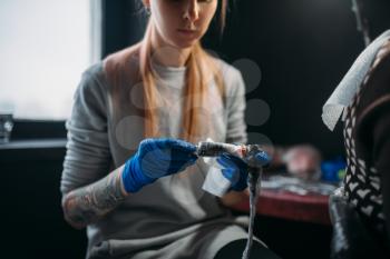Female tattooist in blue sterile gloves prepares tattoo machine, master in salon. Professional tattooing in studio