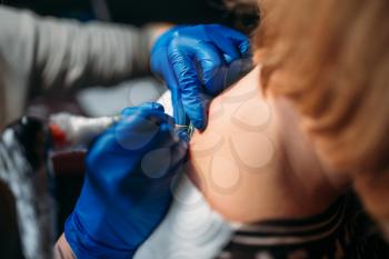 Female tattooer in blue sterile gloves makes tattoo by machine, master in salon. Professional tattooing process in studio