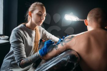 Female tattooist makes tattoo by machine on male shoulder. Professional tattooing in salon